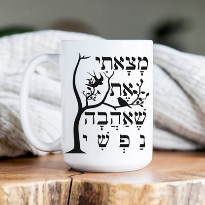 White ceramic mug with the Hebrew phrase "מצאתי את שאהבה נפשי" (I have found the one whom my soul loves) alongside a tree and bird illustration, placed on a wooden surface with a cozy blanket in the background.