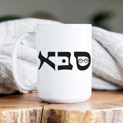 White ceramic mug with the Hebrew word "סבא" (Grandpa) featuring a charming illustration, placed on a wooden surface with a cozy blanket in the background.