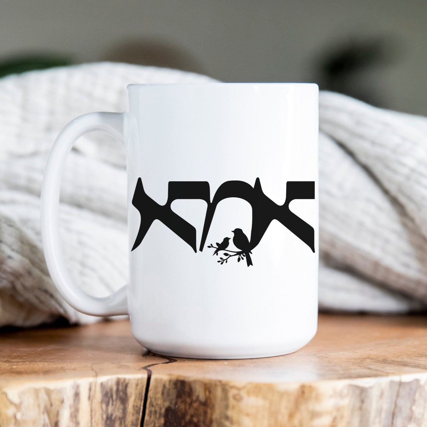 White ceramic mug with the Hebrew word "אמא" (Mom) featuring a bird illustration, placed on a wooden surface with a cozy blanket in the background.