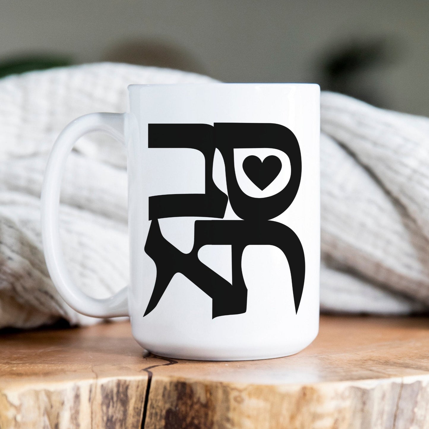 White ceramic mug with the Hebrew word "סבתא" (Grandma) featuring a heart-shaped illustration, placed on a wooden surface with a cozy blanket in the background.