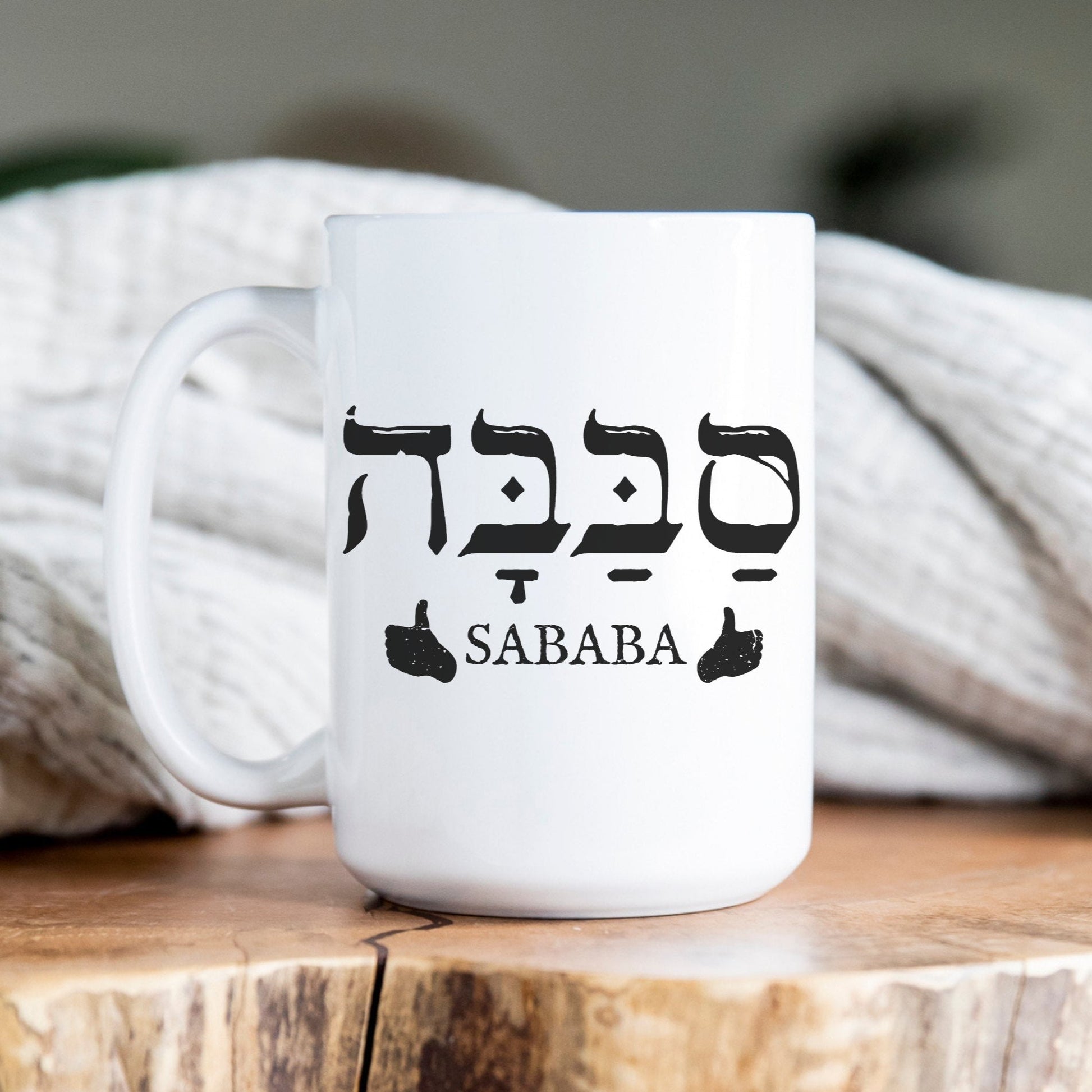 White ceramic mug with the Hebrew word "סבבה" (Sababa) meaning "cool" or "great," and a thumbs-up symbol, placed on a wooden surface with a cozy blanket in the background.