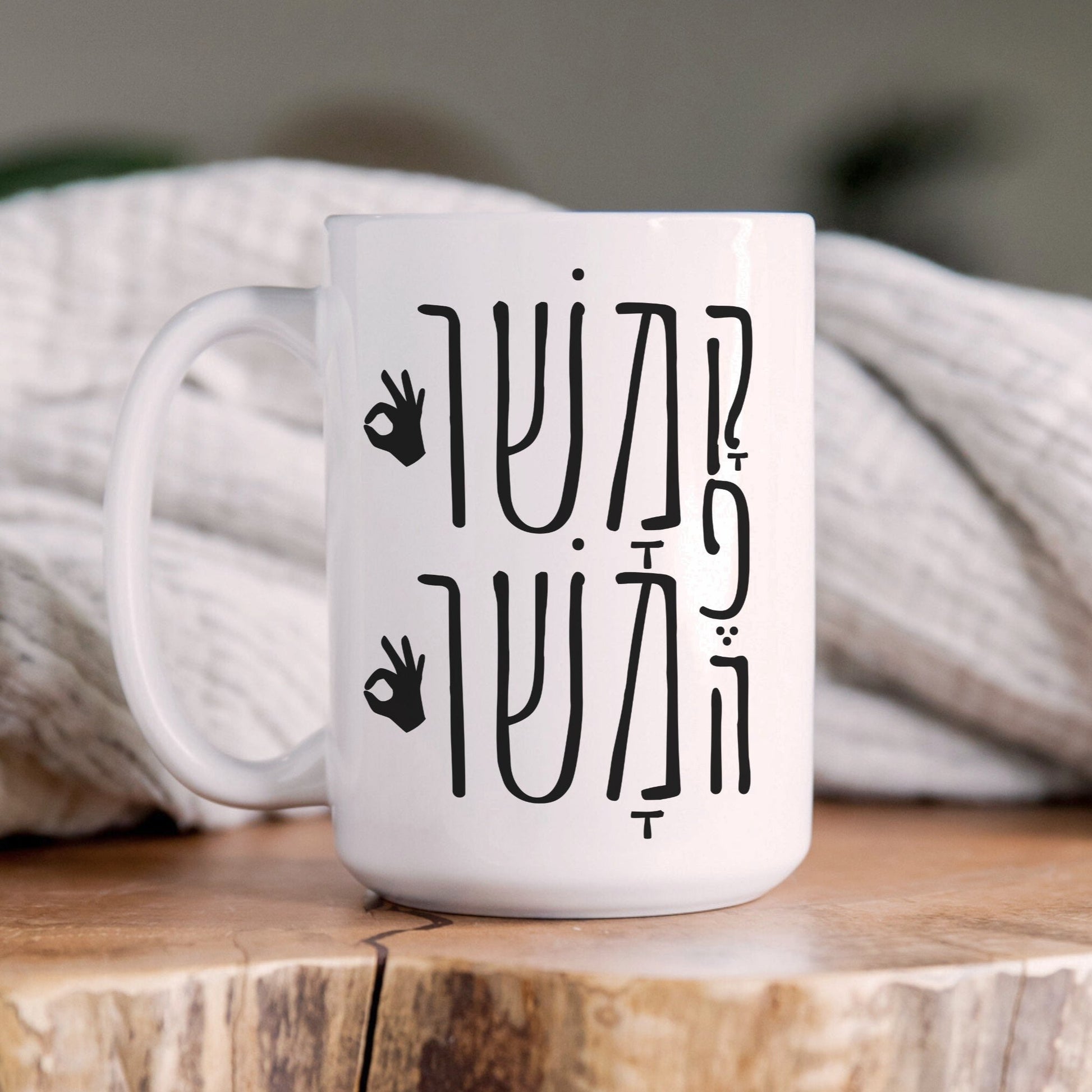 White ceramic mug with "קפה משו משו" (Coffee Mashu Mashu) in Hebrew, featuring "OK" hand signs, placed on a wooden surface with a cozy blanket in the background.