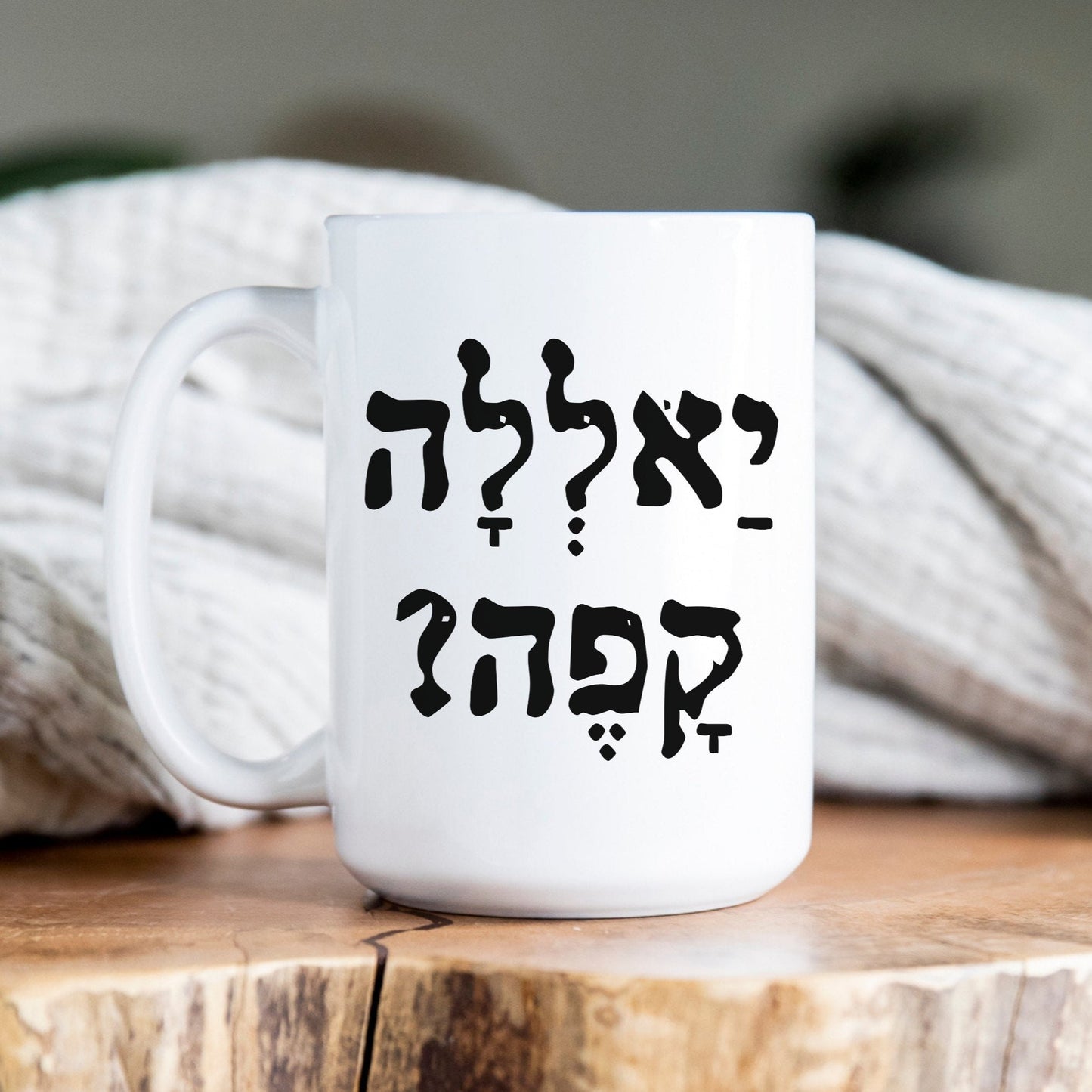 White ceramic mug with the Hebrew phrase "יאללה קפה?" (Yalla, Coffee?), placed on a wooden surface with a cozy blanket in the background.