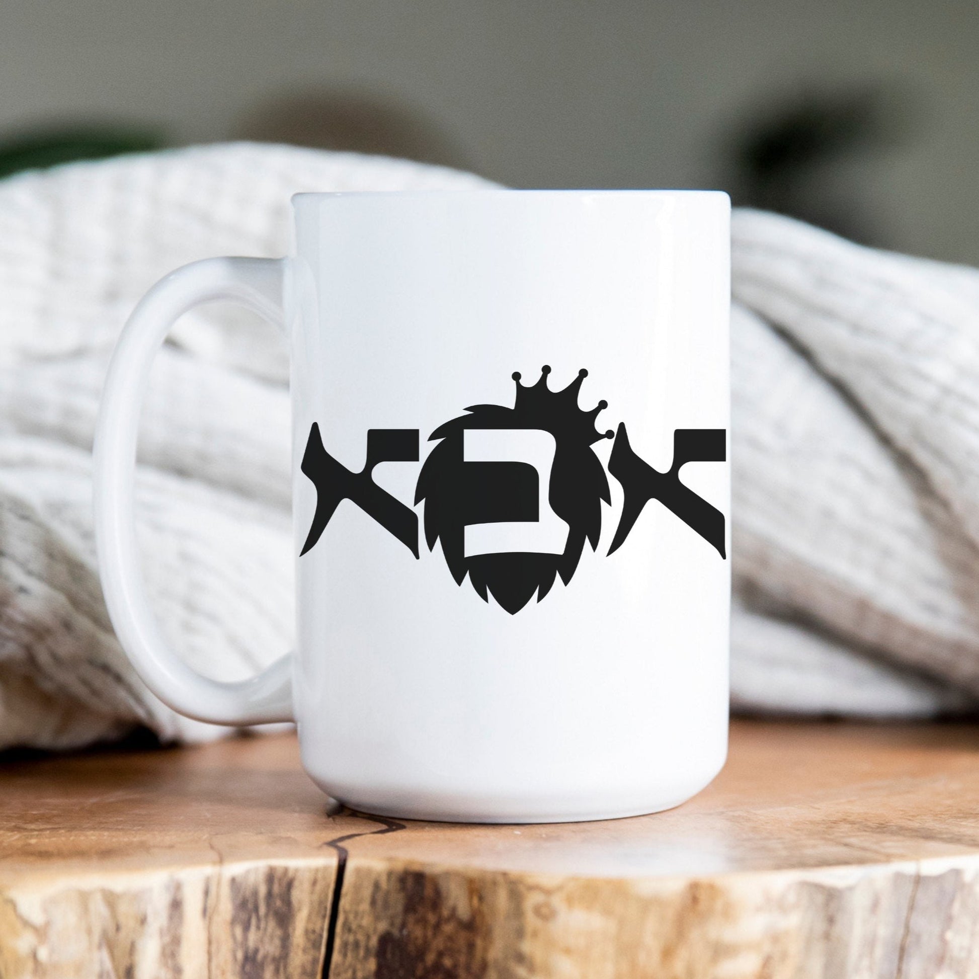 White ceramic mug with the Hebrew word "אבא" (Dad) featuring a crown illustration, placed on a wooden surface with a cozy blanket in the background.