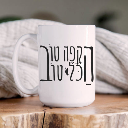 White ceramic mug with the Hebrew phrase "הקפה טוב, הכל טוב" (When the coffee is good, everything is good) and a peace sign, placed on a wooden surface with a cozy blanket in the background.