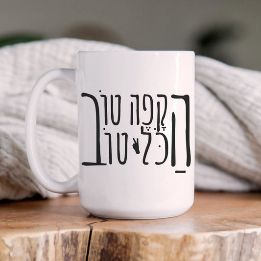 White ceramic mug with the Hebrew phrase "הקפה טוב, הכל טוב" (When the coffee is good, everything is good) and a peace sign, placed on a wooden surface with a cozy blanket in the background.