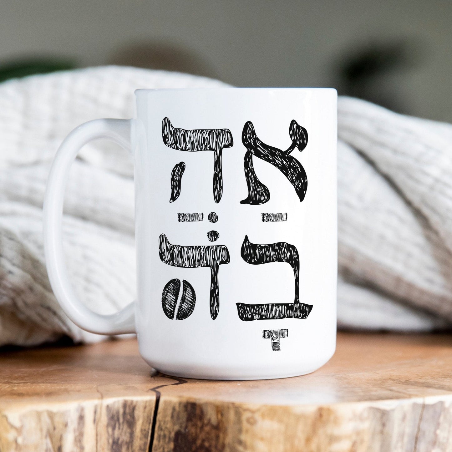 White ceramic mug with the Hebrew word "אהבה" (Love) featuring a coffee bean illustration, placed on a wooden surface with a cozy blanket in the background.