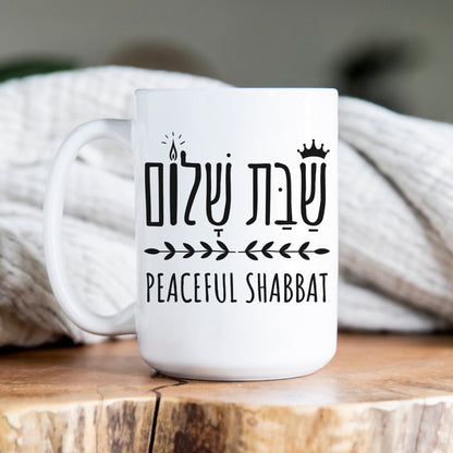 White ceramic mug with the Hebrew phrase "שבת שלום" (Peaceful Shabbat) alongside an illustration of a candle, crown, and olive branch, placed on a wooden surface with a cozy blanket in the background.