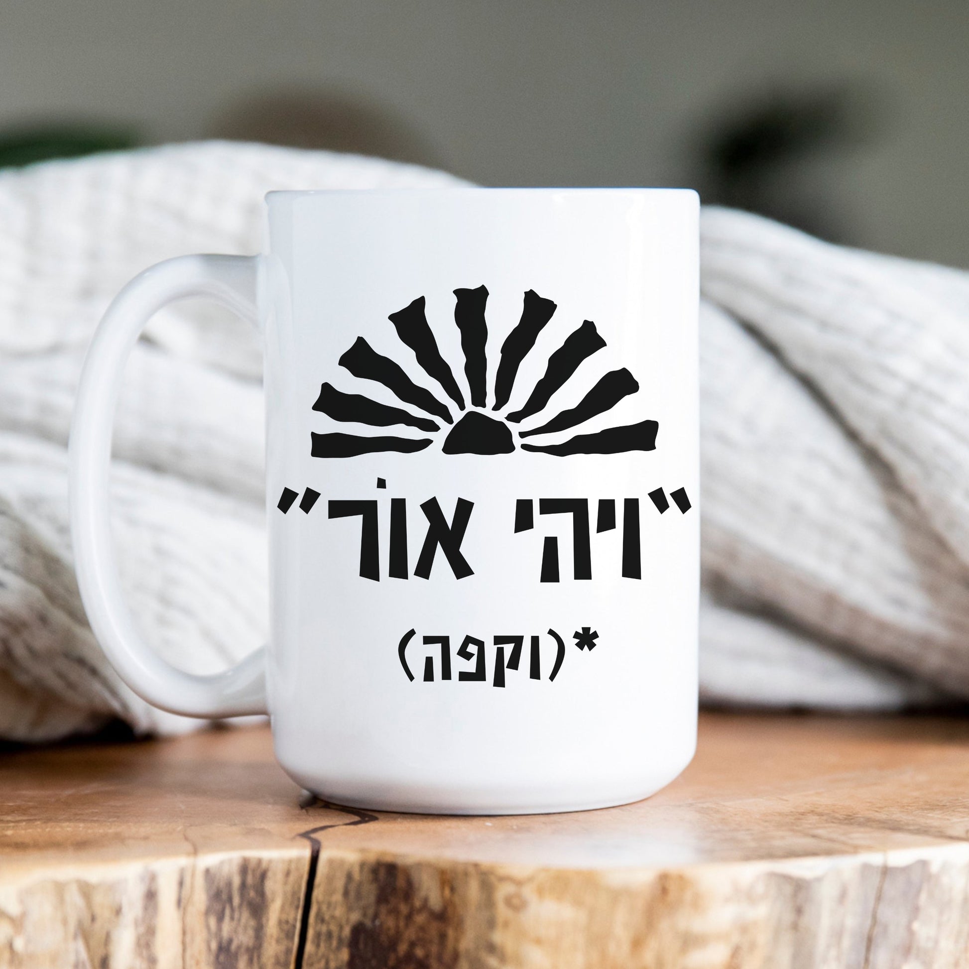 A white ceramic mug featuring the biblical quote "ויהי אור" (Let There Be Light) with the humorous addition "וקפה" (and coffee) and a sun illustration, sitting on a wooden plate with a cozy blanket behind it.