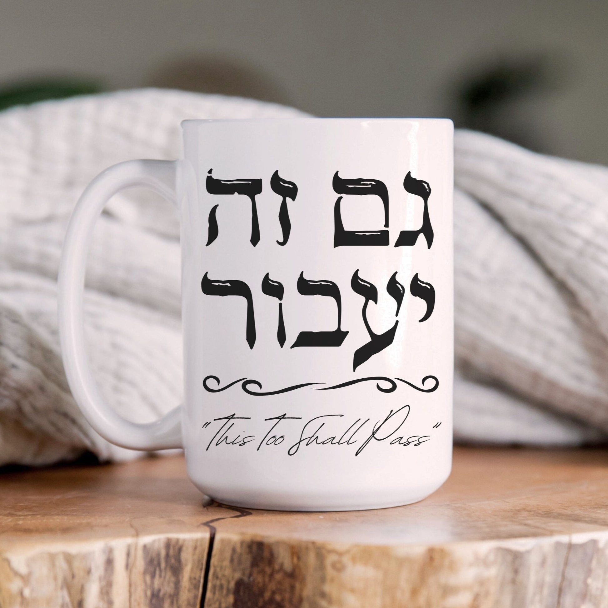 A white ceramic mug featuring the Hebrew phrase "גם זה יעבור" (This Too Shall Pass) in black text, sitting on a wooden surface with a cozy blanket in the background.