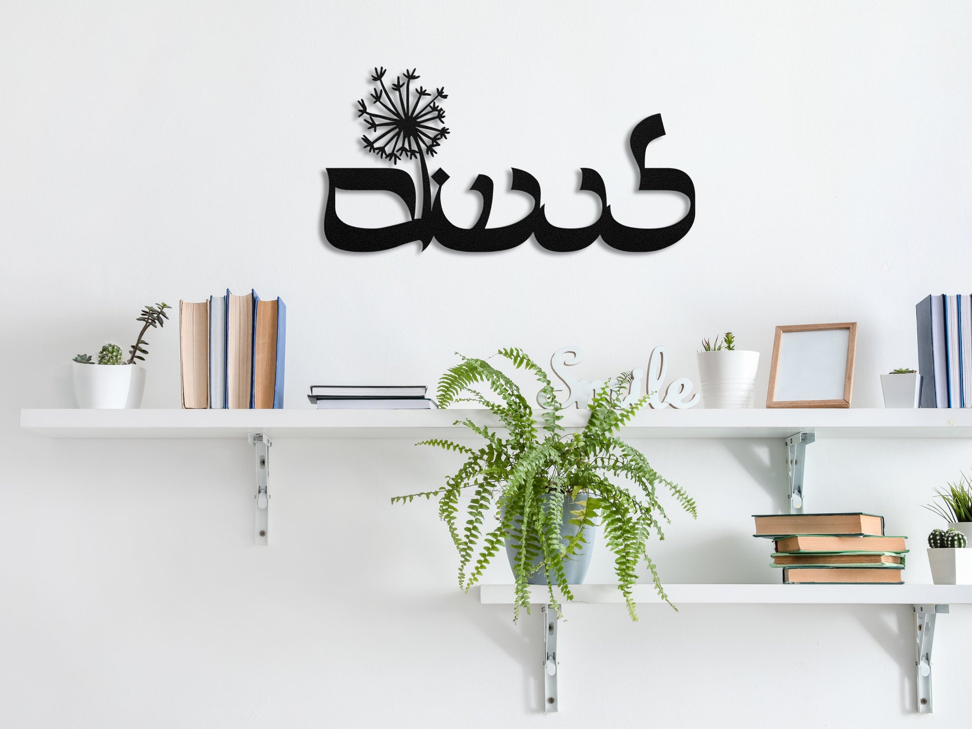 Black metal sign of the word "לנשום" with a dandelion, hanging on a wall above white shelves decorated with books and plants.