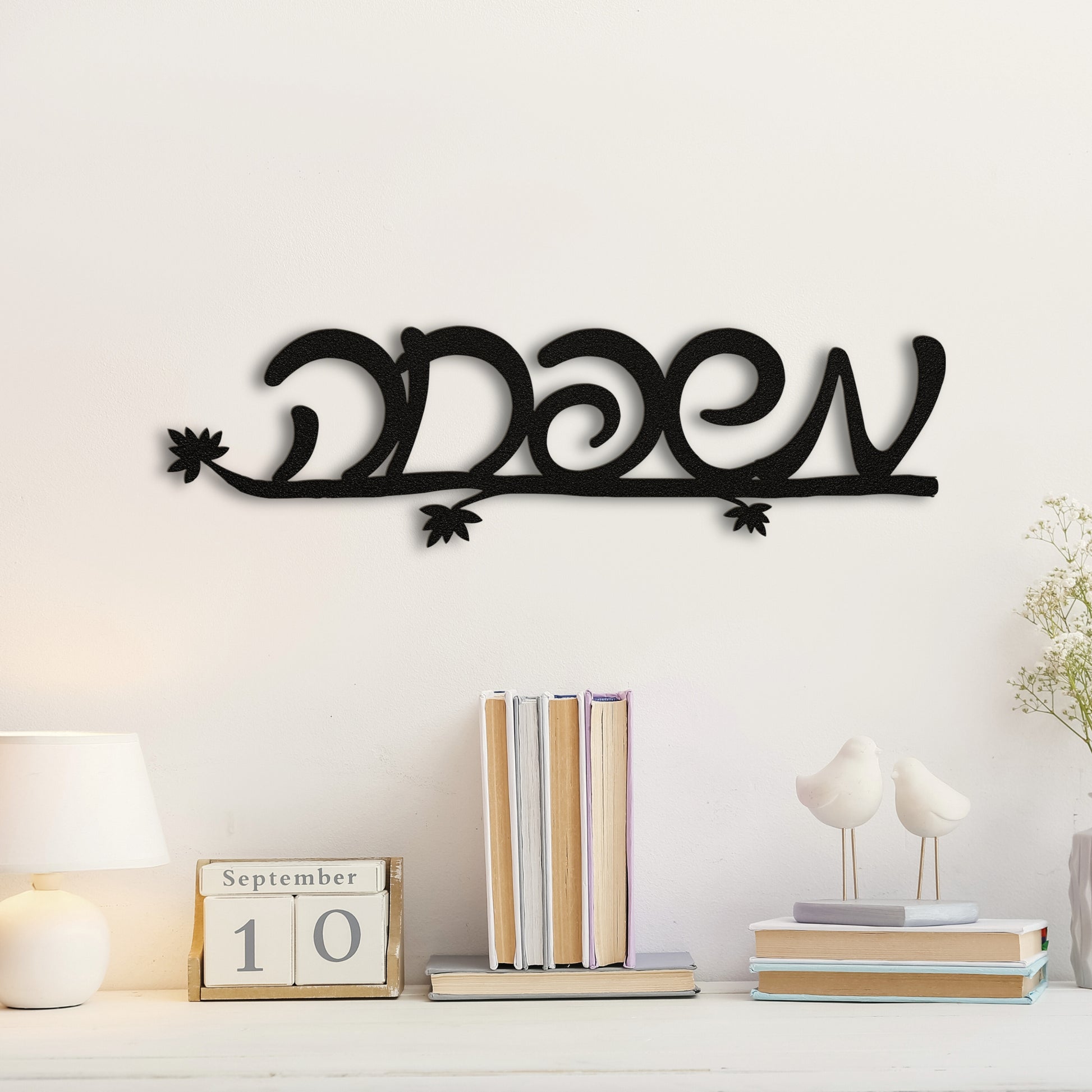 Black metal wall art with the Hebrew word 'משפחה' (Family) mounted on a white wall above a white desk with books and decor items.