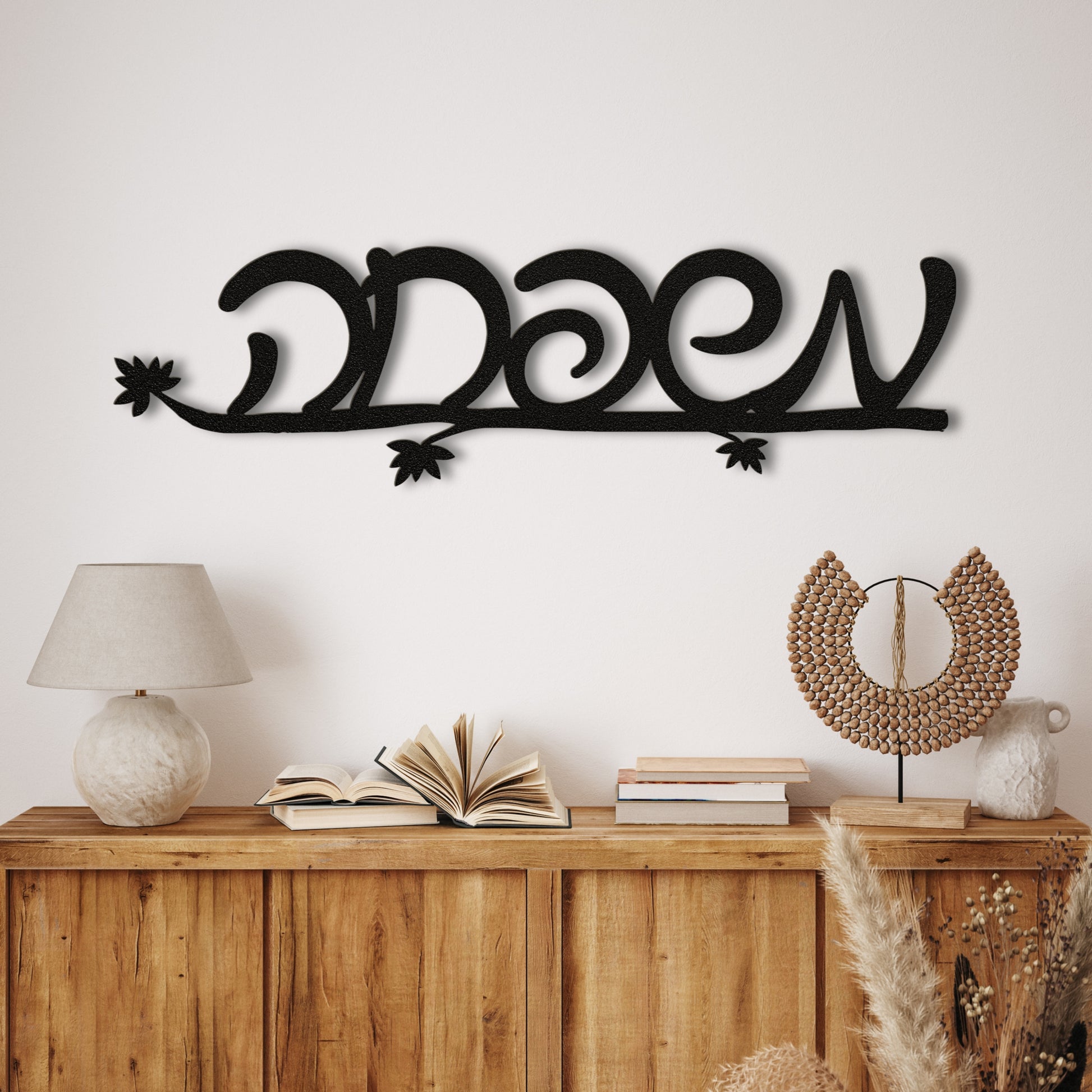 Black metal wall art with the Hebrew word 'משפחה' (Family) on a white wall above a wooden shelf with decor items.