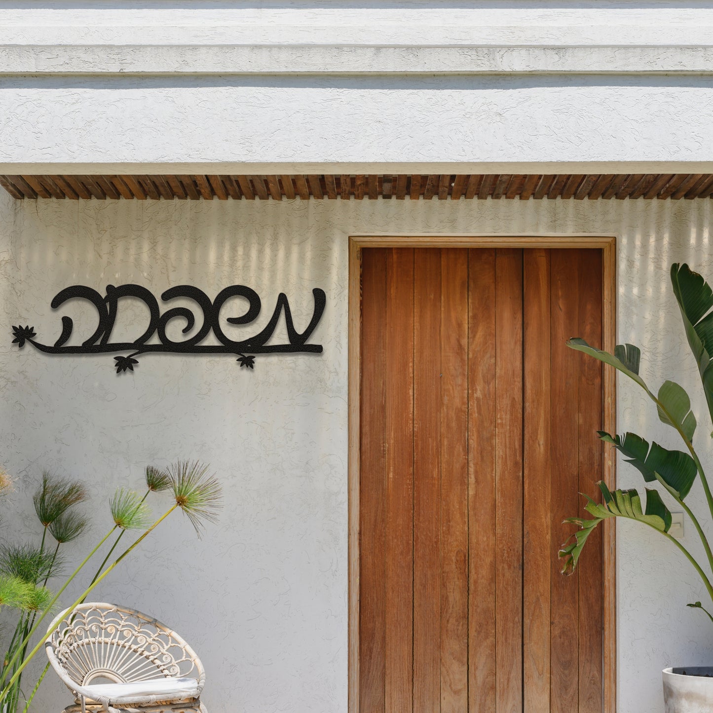 Black metal wall art with the Hebrew word 'משפחה' (Family) mounted outside above a wooden door, with plants nearby.