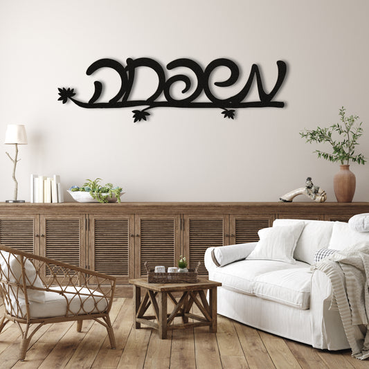 Black metal wall art with the Hebrew word 'משפחה' (Family) on a light beige wall above a wooden sideboard with a cozy living room setup.