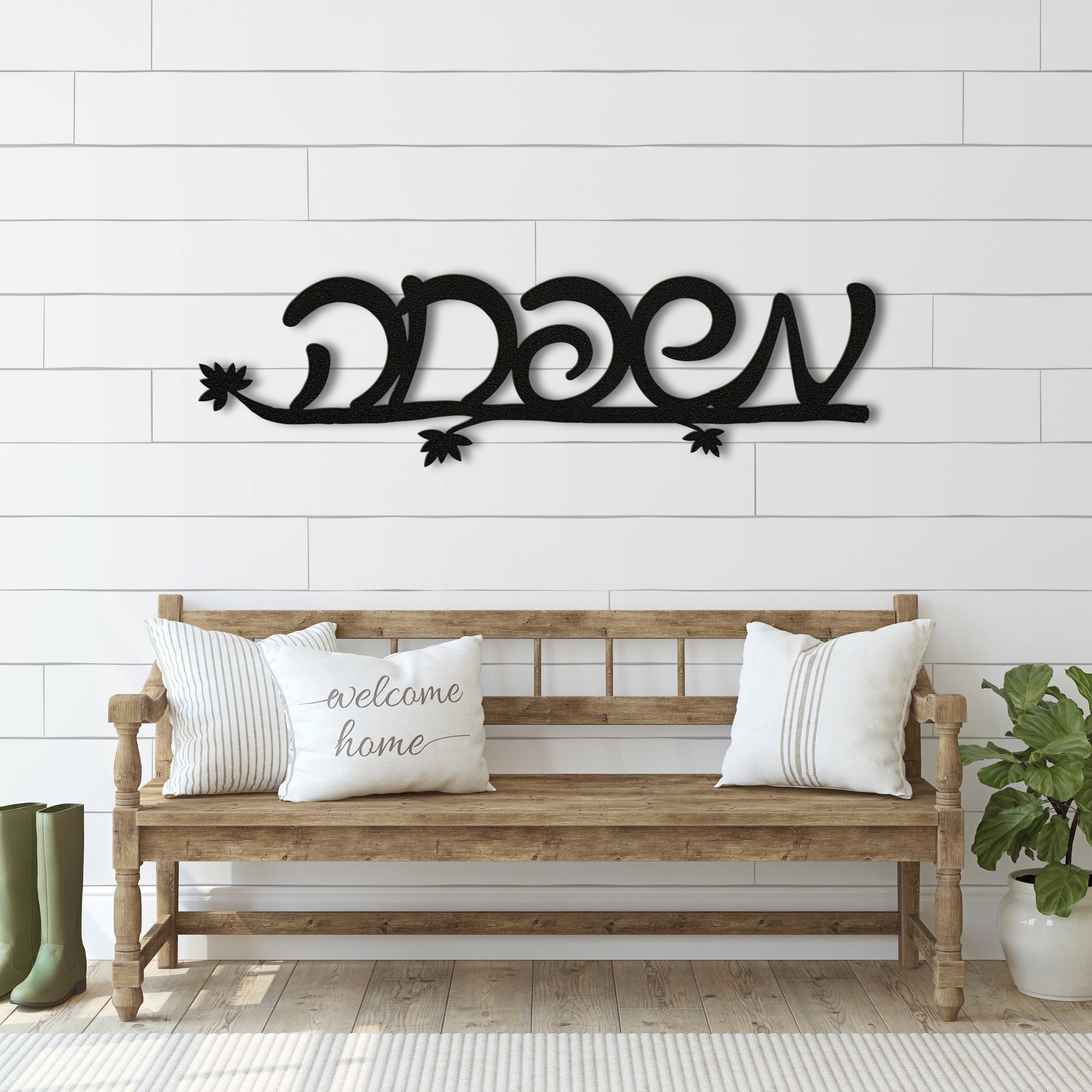 Black metal wall art with the Hebrew word 'משפחה' (Family) on a white shiplap wall above a wooden bench with decorative pillows.