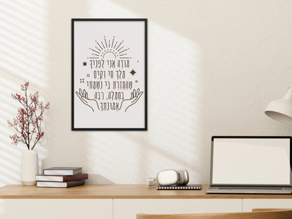 Framed wall art with the Hebrew phrase 'מודה אני' (Modeh Ani), featuring hands and sunburst design, hung above a desk with a lamp and books.