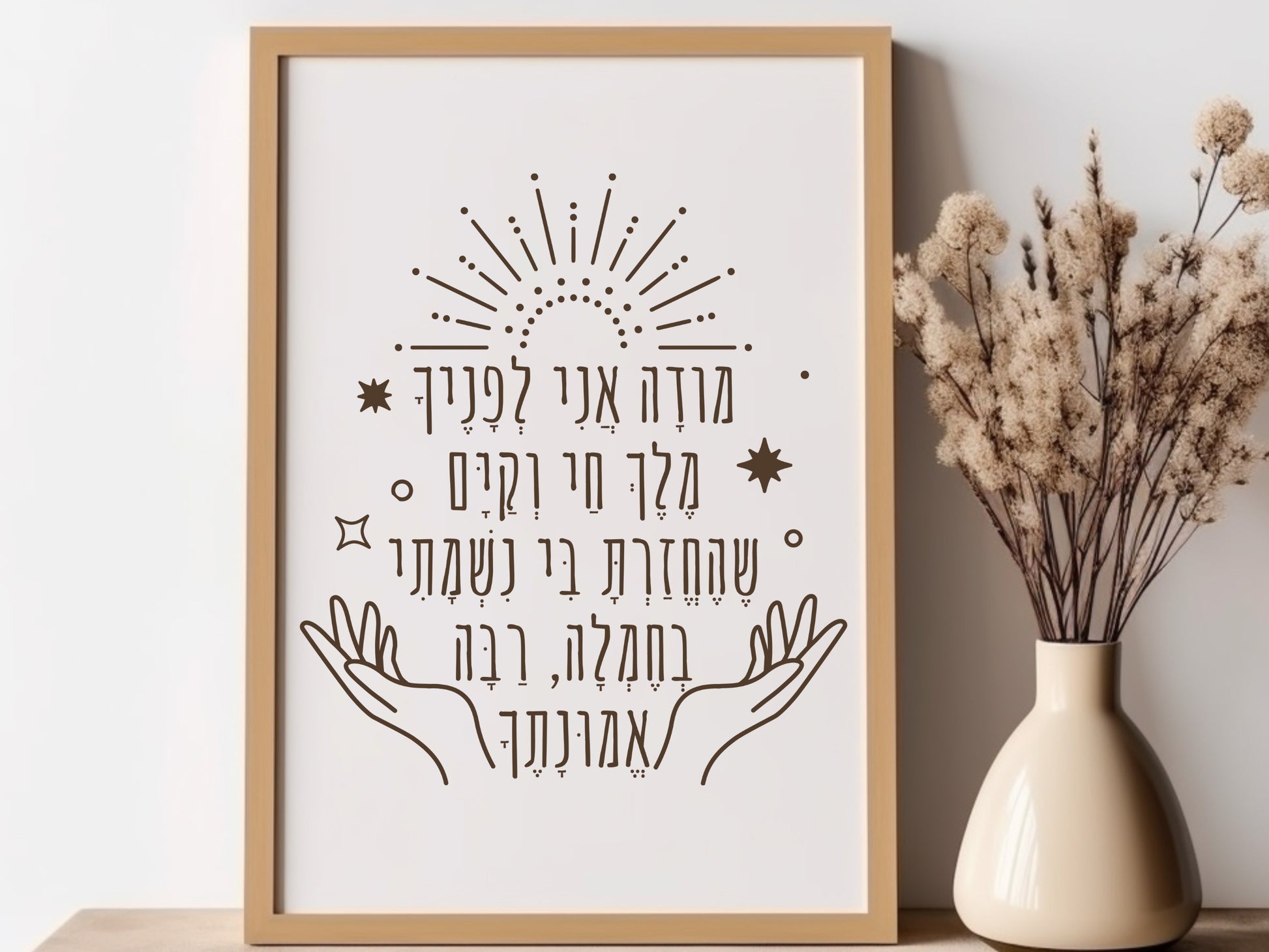 Framed wall art with the Hebrew phrase 'מודה אני' (Modeh Ani), featuring hands and sunburst design, placed next to a vase with dried flowers.