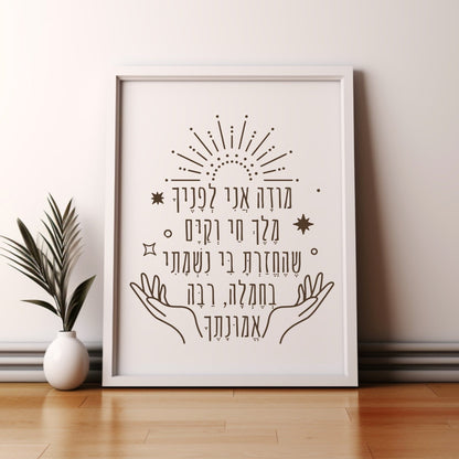 Framed wall art with the Hebrew phrase 'מודה אני' (Modeh Ani), featuring hands and sunburst design, leaning against a wall beside a small potted plant.