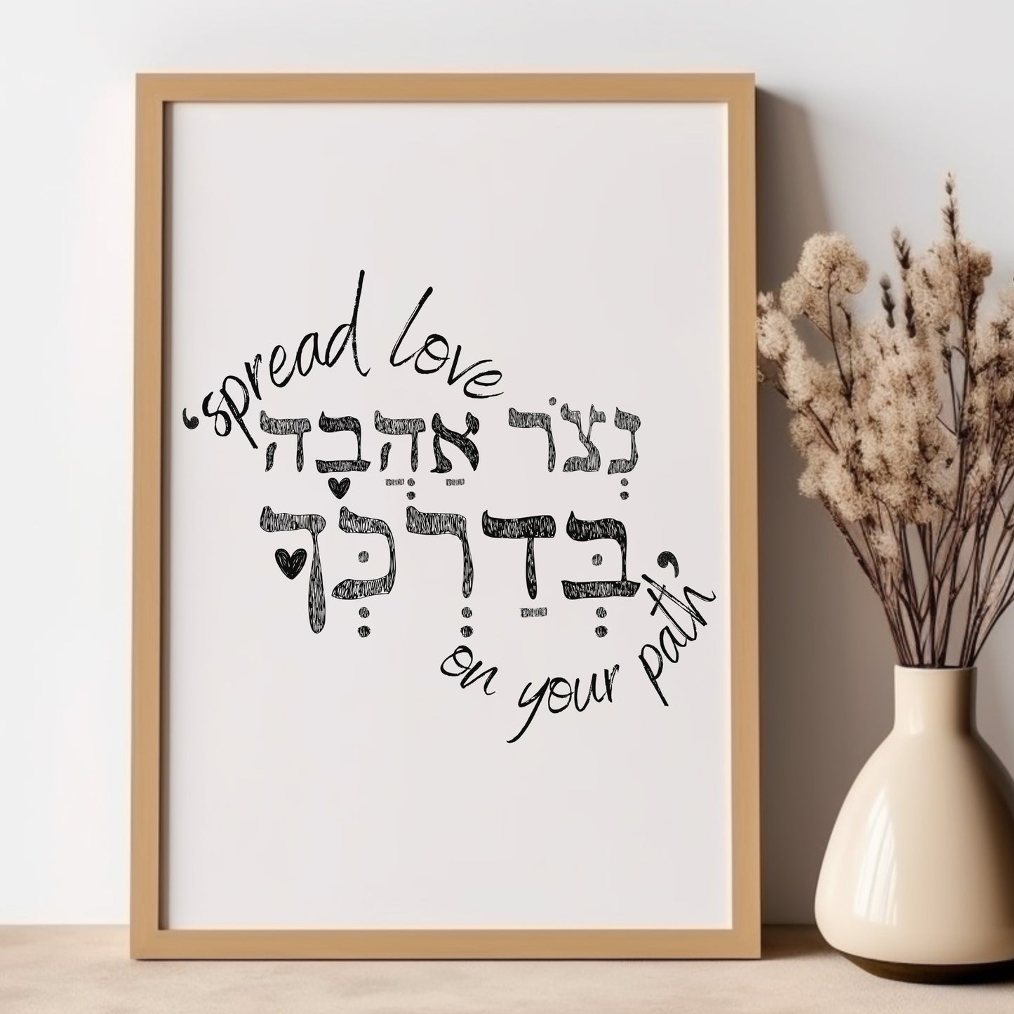 Framed wall art with the Hebrew phrase 'נצור אהבה בדרכך' (Spread love on your path), featuring a minimalist design, placed next to a vase with dried flowers.