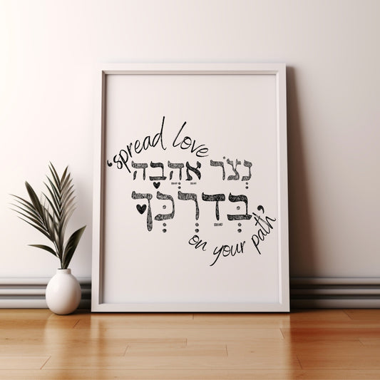 Framed wall art with the Hebrew phrase 'נצור אהבה בדרכך' (Spread love on your path), featuring a minimalist design, leaning against a wall beside a small potted plant.