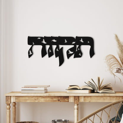 Metal wall art with the word 'תקווה' (Hope) in Hebrew, intricately designed with dove silhouettes, displayed on a white wall above a wooden console table with books and dried floral arrangements.