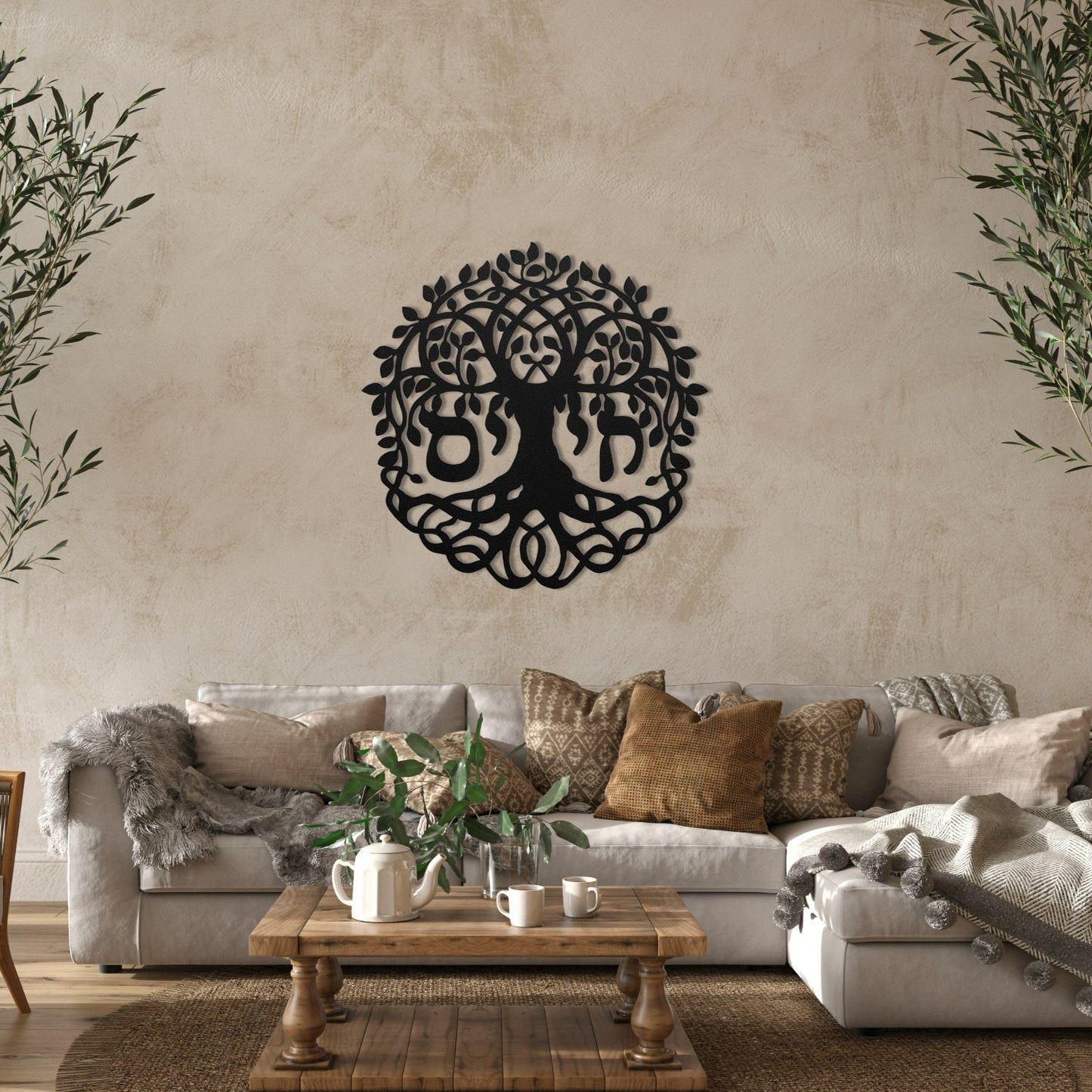 A beautiful metal wall art featuring the Hebrew word "חי" (Chai) meaning "Life," incorporated into a detailed Tree of Life design. The artwork is displayed in a cozy, rustic living room with a beige sofa, earth-toned cushions, and a wooden coffee table adorned with plants and tea set.