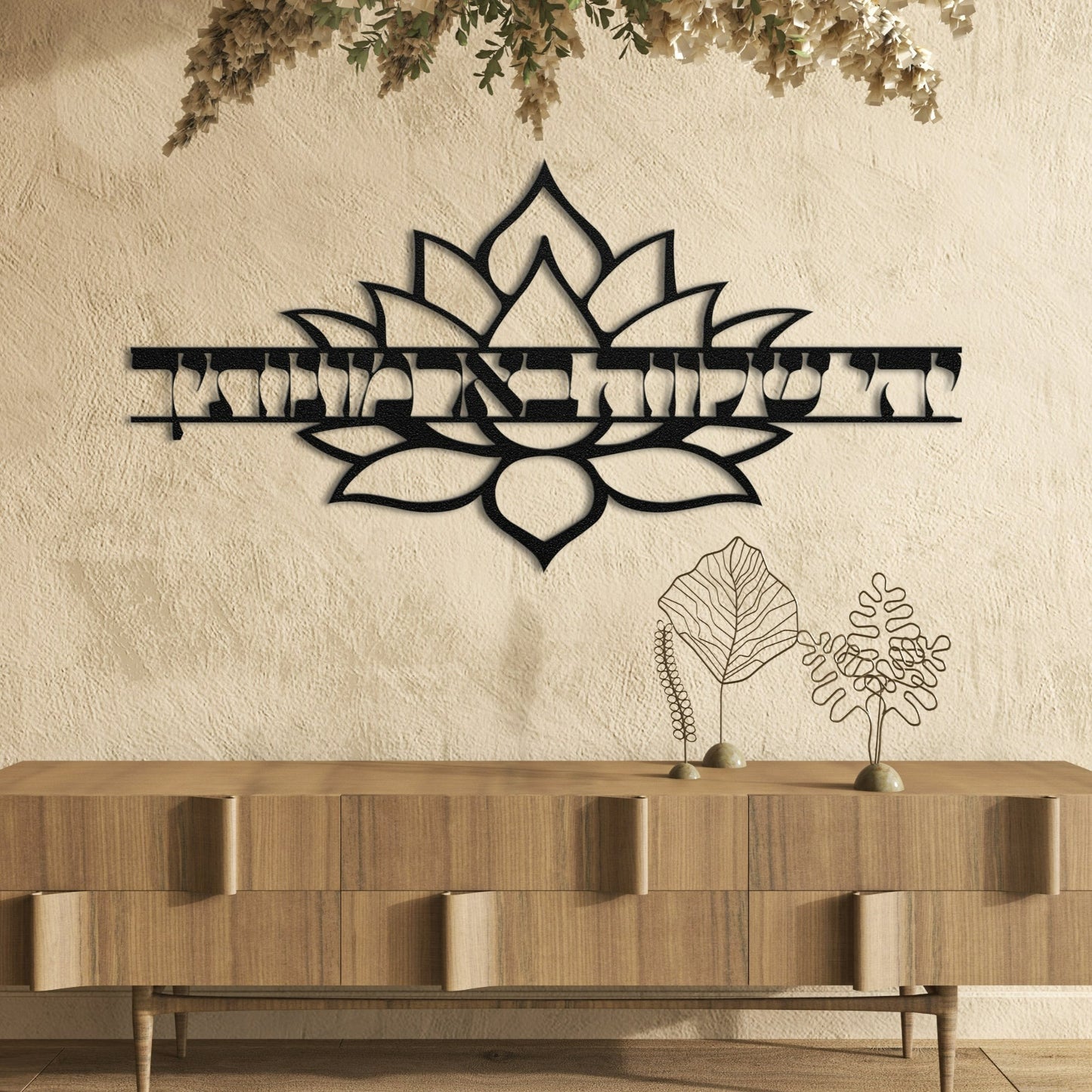 A stylish metal wall art piece featuring the Hebrew phrase "יהי שלווה בארמונותיך" (May there be peace within your walls) with a decorative lotus flower design. The art is displayed against a textured wall above a wooden cabinet, creating a harmonious and serene ambiance.