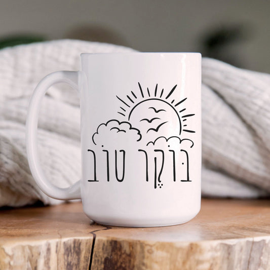 White ceramic mug with "בוקר טוב" (Good Morning) in Hebrew, featuring an illustration of the sun, clouds, and birds, placed on a wooden surface with a cozy blanket in the background.