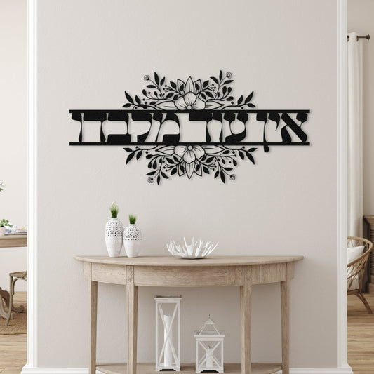 A stylish entryway with a wooden console table decorated with white vases and seashells. Above the table, there is a black metal wall art piece with the Hebrew words "אין עוד מלבדו" (None but Him) surrounded by an intricate floral design.