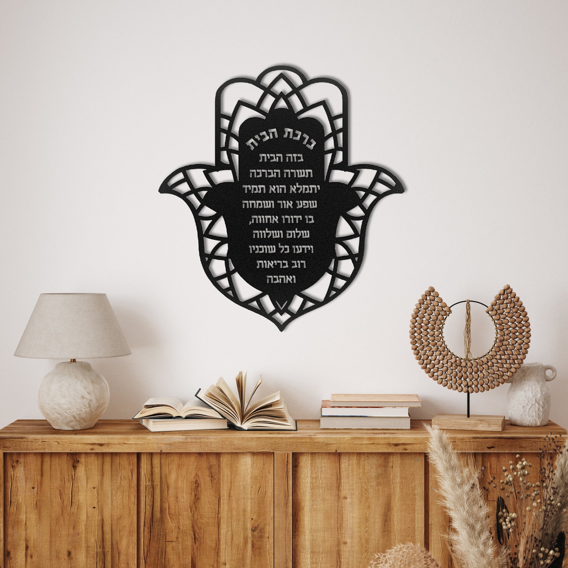 A beautifully designed metal wall art in the shape of a Hamsa, featuring the "Birkat Habayit" (Jewish Home Blessing) in Hebrew. The artwork is intricately crafted with a floral pattern surrounding the blessing text. It is displayed on a light-colored wall above a wooden console table with decorative items, creating an elegant and serene atmosphere in the room.