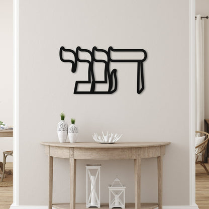Elegant metal wall art featuring the Hebrew word 'הנני' (here I am), displayed in a serene, modern living space with minimalistic decor and neutral tones.