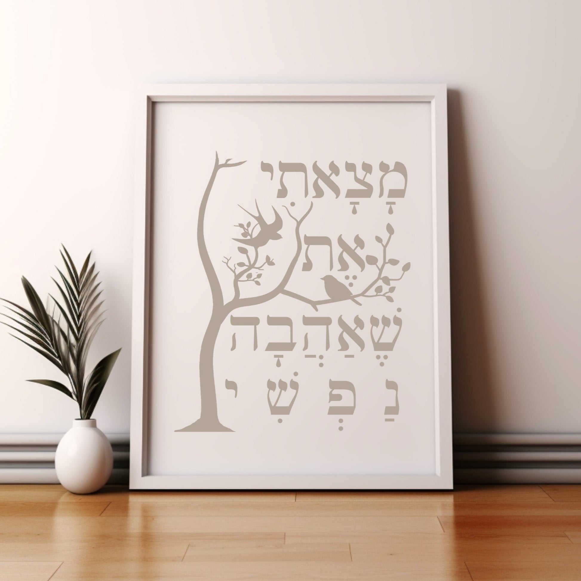 Framed wall art featuring a tree and the Hebrew verse 'מצאתי את שאהבה נפשי' (I have found the one whom my soul loves), leaning against a wall next to a small potted plant.