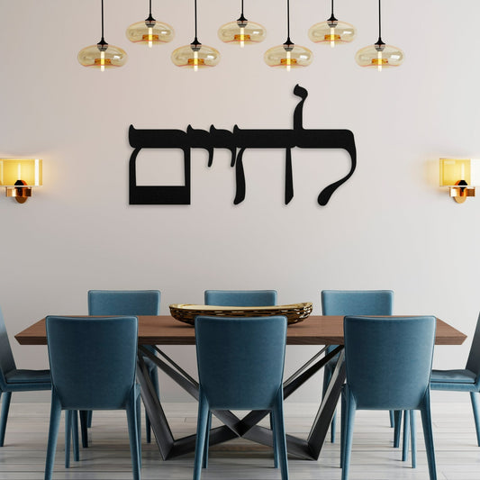 A stylish "לחיים" (To Life) metal wall art piece hangs above a modern dining table set with blue chairs, framed by warm yellow wall sconces, creating a welcoming and elegant atmosphere.