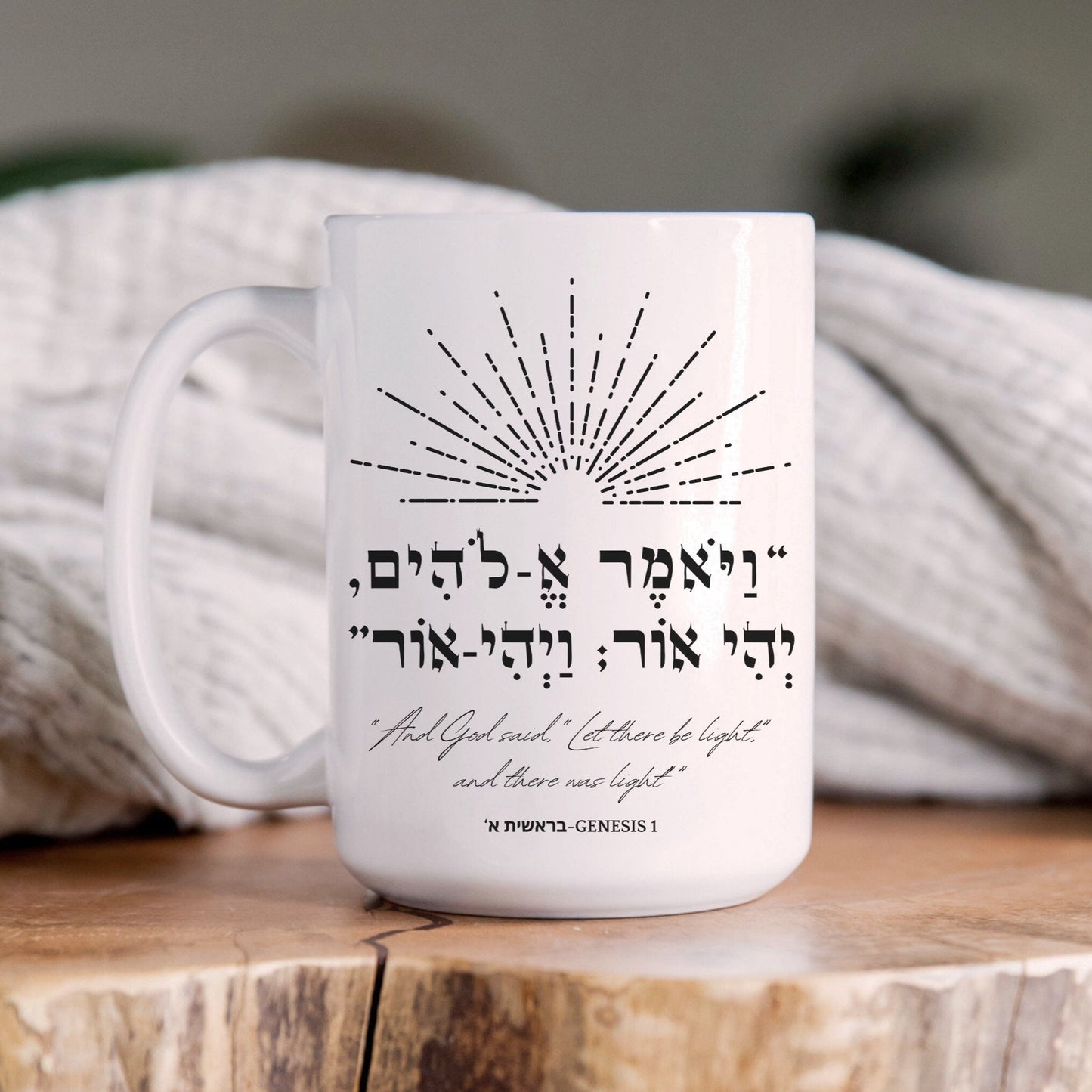 A white ceramic mug featuring the biblical Hebrew verse "ויאמר אלהים יהי אור ויהי אור" (And God said, 'Let there be light'; and there was light) with an illustration of light rays, placed on a wooden surface with a cozy blanket in the background.