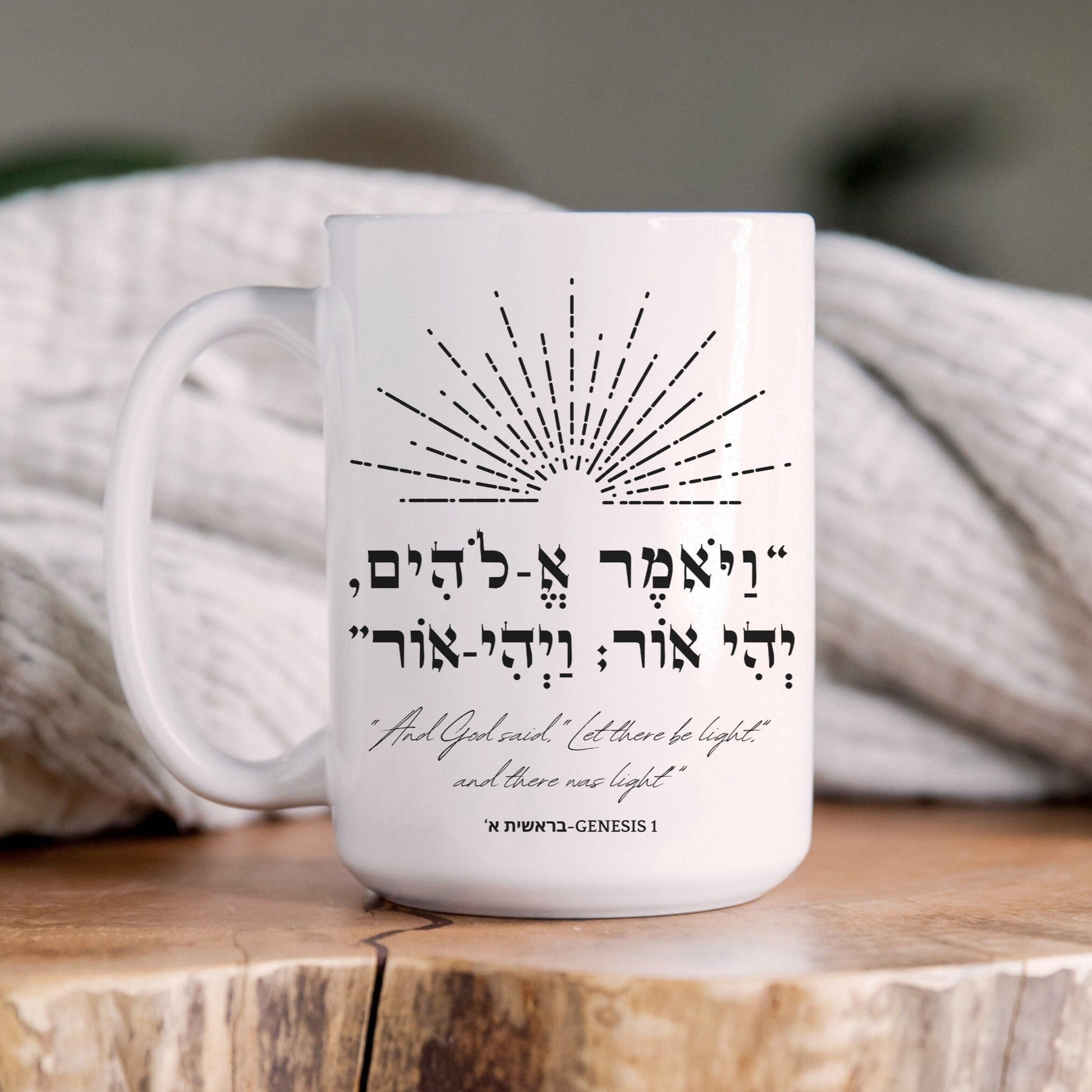 A white ceramic mug featuring the biblical Hebrew verse "ויאמר אלהים יהי אור ויהי אור" (And God said, 'Let there be light'; and there was light) with an illustration of light rays, placed on a wooden surface with a cozy blanket in the background.