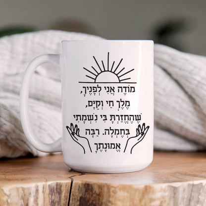 "מודה אני" Hebrew mug featuring the Jewish morning blessing, with a sun illustration above the text, placed on a wooden surface with a cozy blanket in the background.