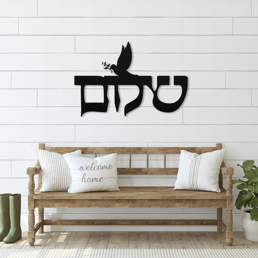 Metal wall art featuring the Hebrew word 'שלום' (Shalom) meaning peace, with a dove carrying an olive branch. The sign is displayed above a bench in a bright entryway with plants and a 'welcome home' pillow.