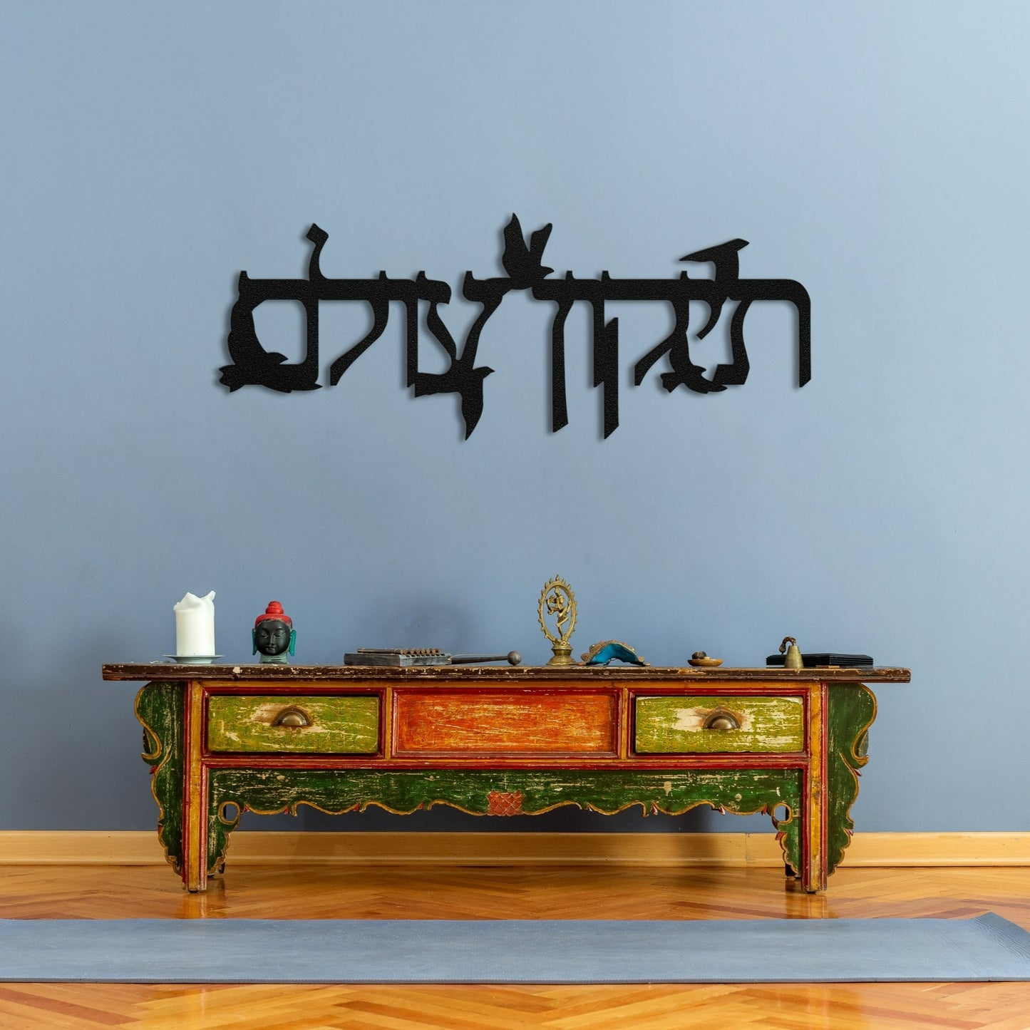 Metal sign "תיקון עולם" with flying doves featured in a tranquil yoga space, positioned above an antique table with colorful decor and a yoga mat.