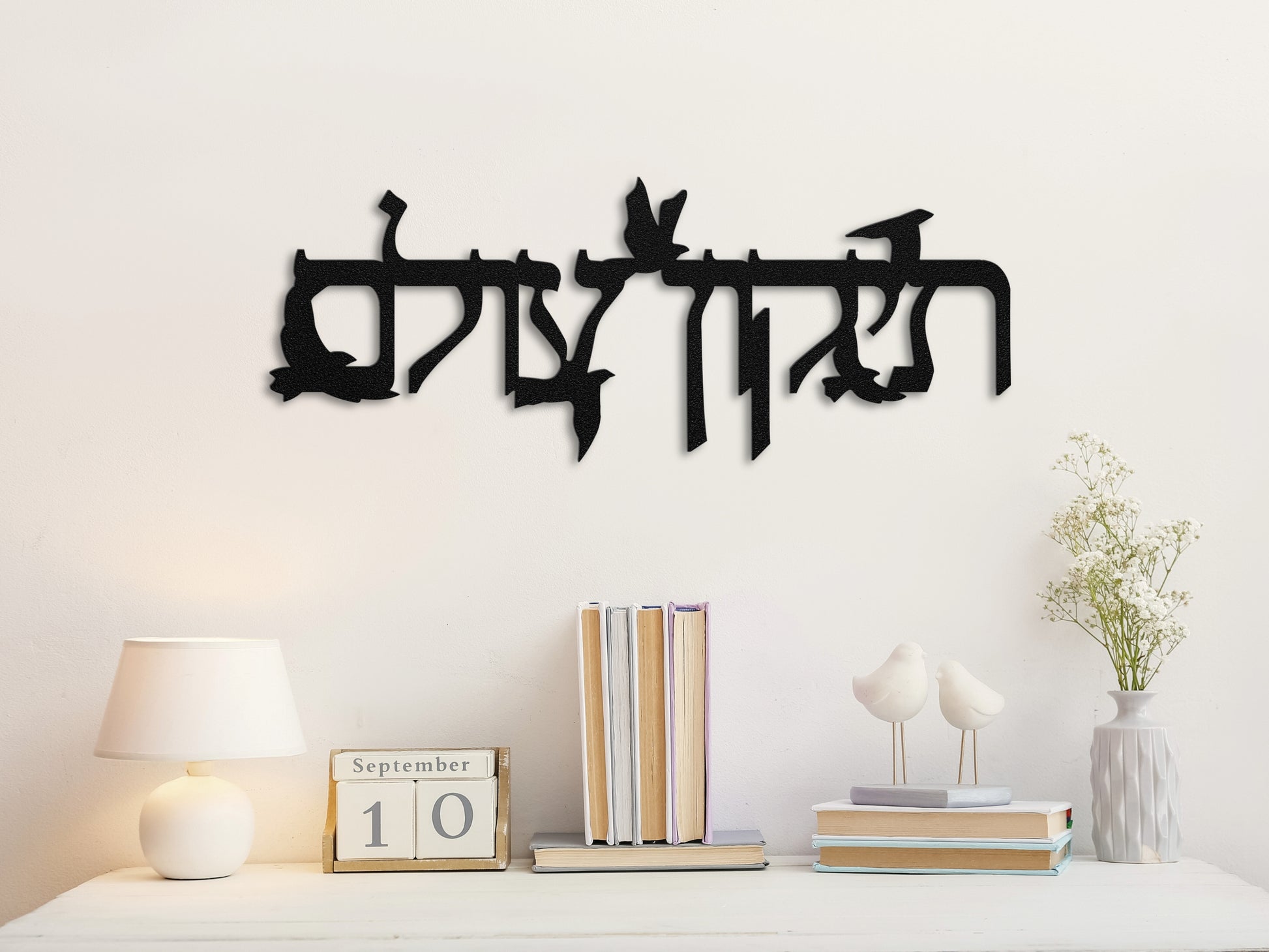 Metal sign "תיקון עולם" with flying doves displayed above a neat bookshelf with books, decor items, and a minimalist, peaceful vibe.